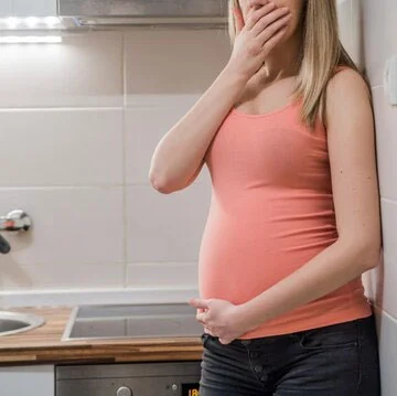 Pregnant woman experiencing nausea and vomiting in the kitchen, with one hand covering her mouth and the other supporting her belly, symbolizing symptoms of nausea and vomiting in pregnancy.