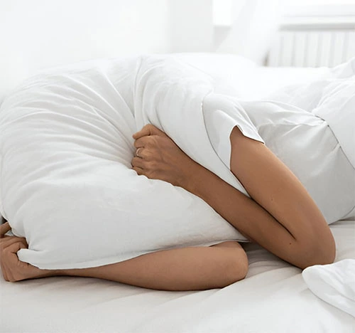 Person sitting on a bed holding their lower back, indicating back pain or discomfort.